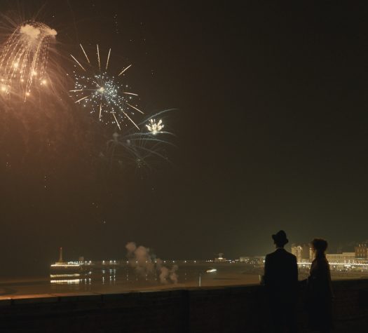 Szeroka panorama miasteczka portowego. Dwie osoby, częściowo zacienione, oglądają pokaz fajerwerków. Obok nich neon z napisem "Empire".