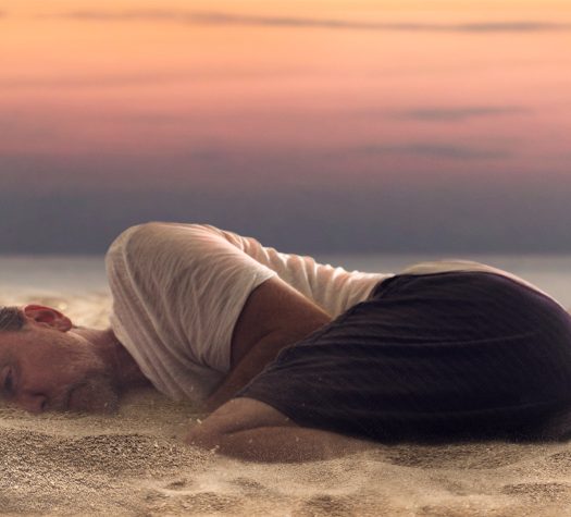 Na zdjęciu mężczyzna leżący na plaży, częściowo zanurzony w piasku. W tle złoto-amarantowe niebo w porze zachodu słońca.