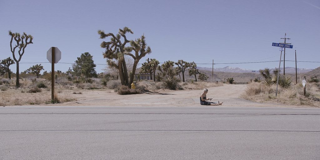 Kobieta siedząca na skraju amerykańskiej drogi na pustkowiu. Kobieta jes bosa. W tle widoczne kaktusy, krajobraz palony intensywnym słońcem.