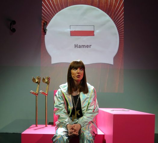 Młoda kobieta ubrana w błyszczący dres, siedzi na różowych kubikach. Za nią kule ortopedyczne koloru złotego. W tle napis „Hamer” i polska flaga.