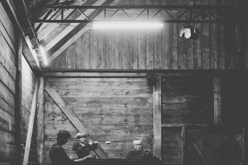 Na czarno-białym zdjęciu trzy osoby, dwóch mężczyzn i jedna kobieta, stoją na tle drewnianej ściany. Wydają się grać na instrumentach, które są niewidoczne.