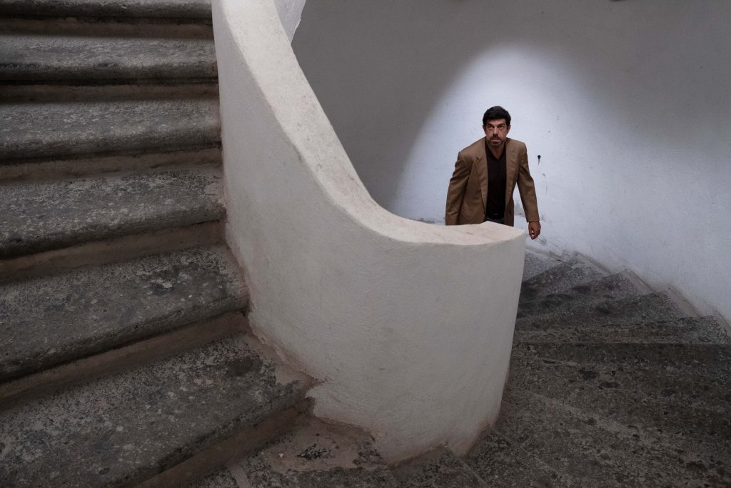Mężczyzna wchodzący po krętych, kamiennych schodach. Kamienna poręcz schodów i ściana za osobą pomalowane na biało.